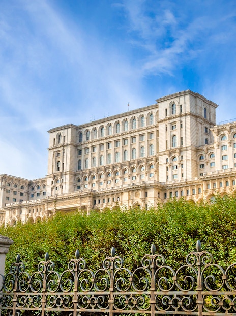 Palace of the Parliament in Bucharest, Romania, with ornate iron fence and lush greenery.