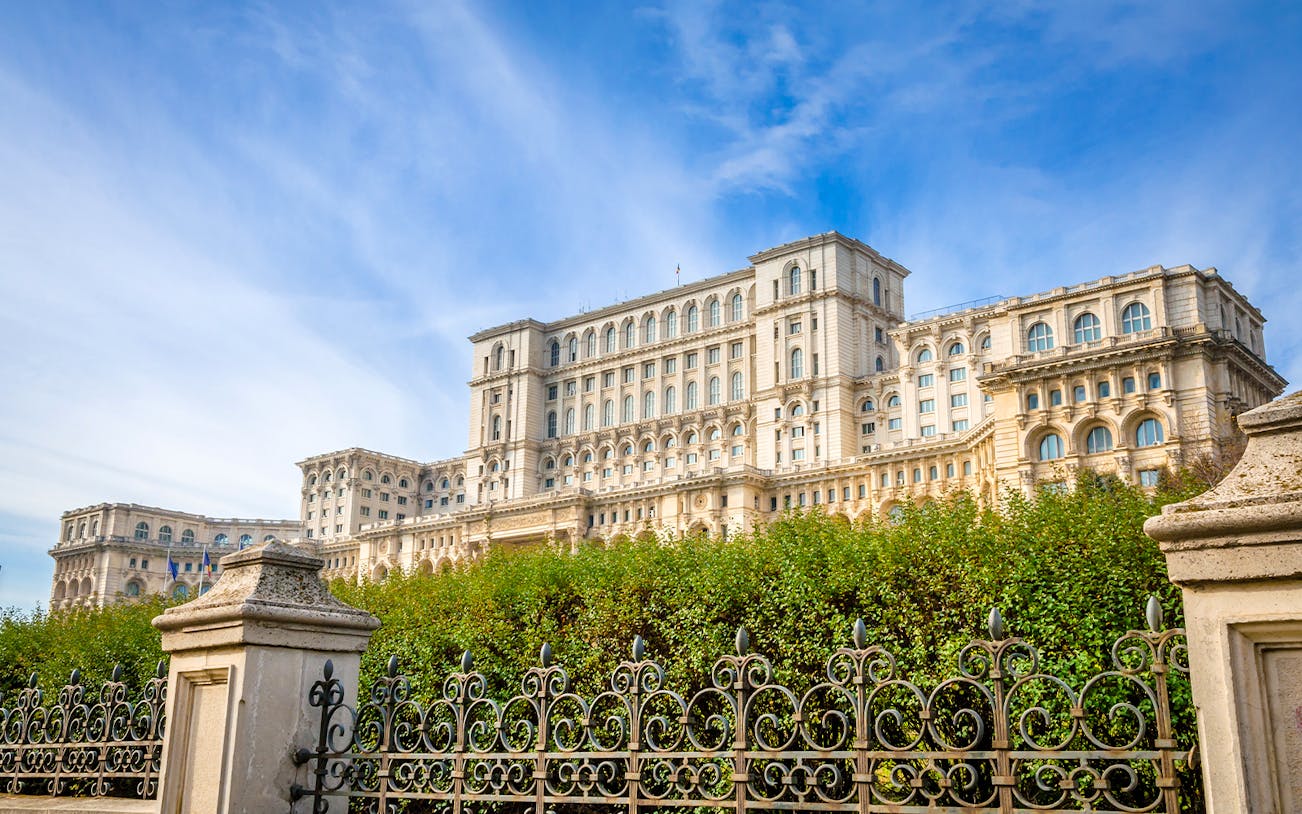 Palace of the Parliament in Bucharest, Romania, with ornate iron fence and lush greenery.