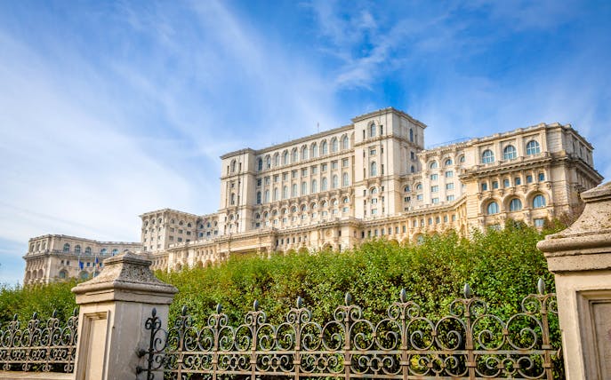 Palace of the Parliament in Bucharest, Romania, with ornate iron fence and lush greenery.