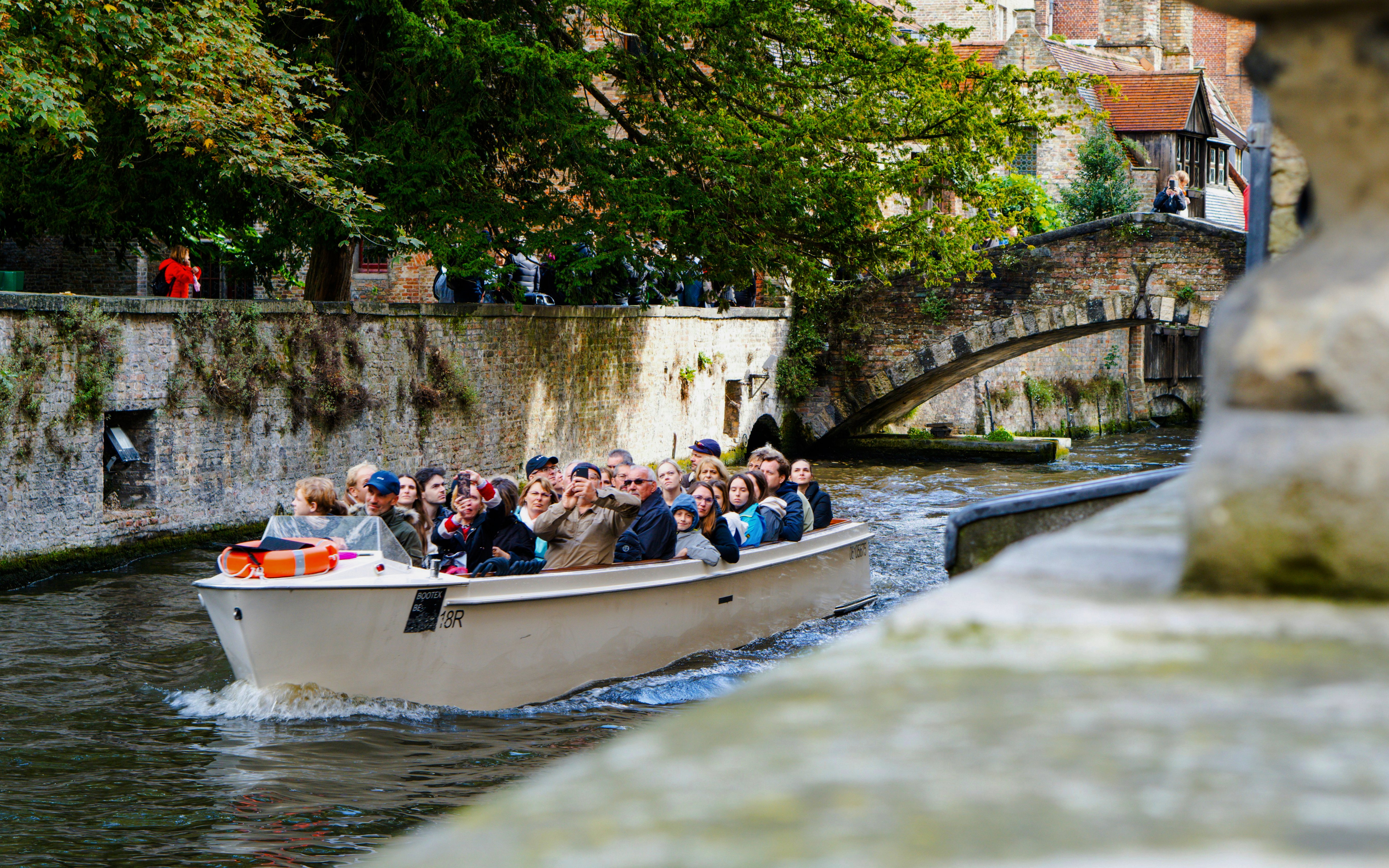Canal boat tour with tourists passing under a stone bridge in Bruges, Belgium.