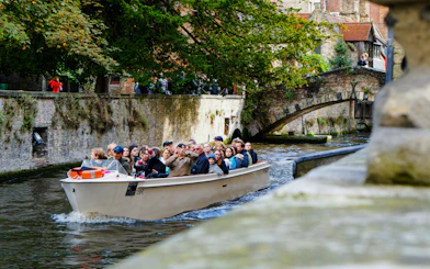 Canal boat tour with tourists passing under a stone bridge in Bruges, Belgium.