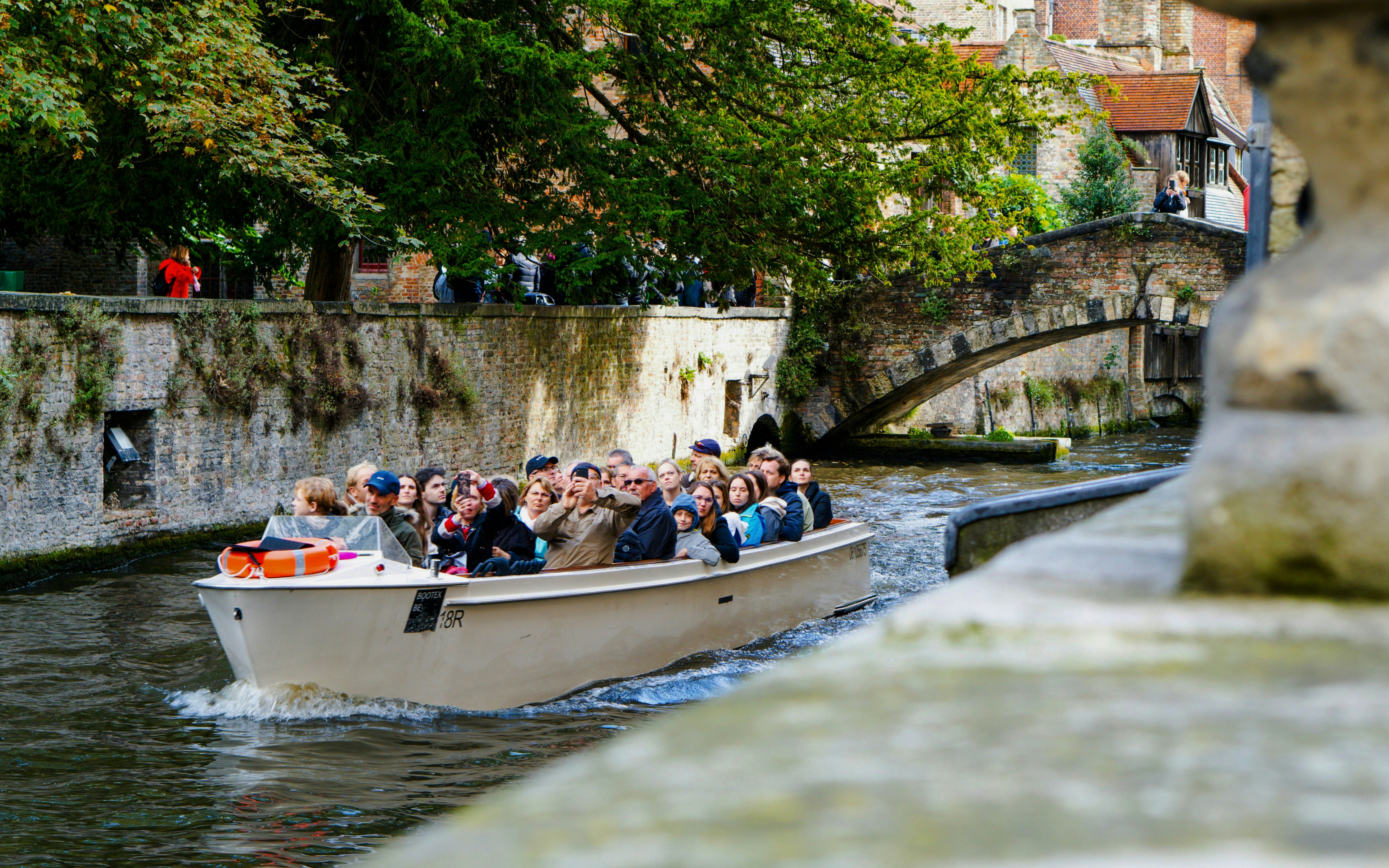 Canal boat tour with tourists passing under a stone bridge in Bruges, Belgium.