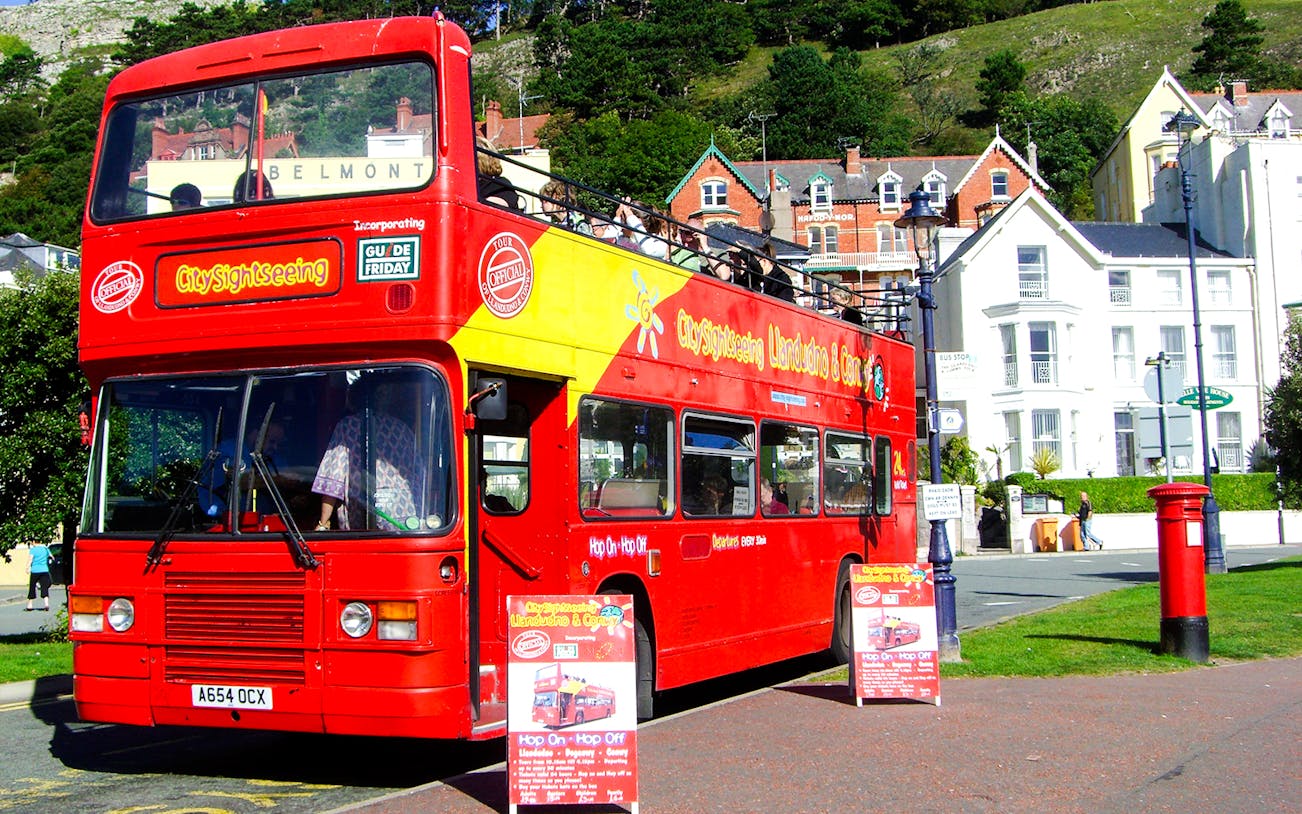 Open-top red sightseeing bus in Llandudno, Wales, with tourists viewing townhouses.