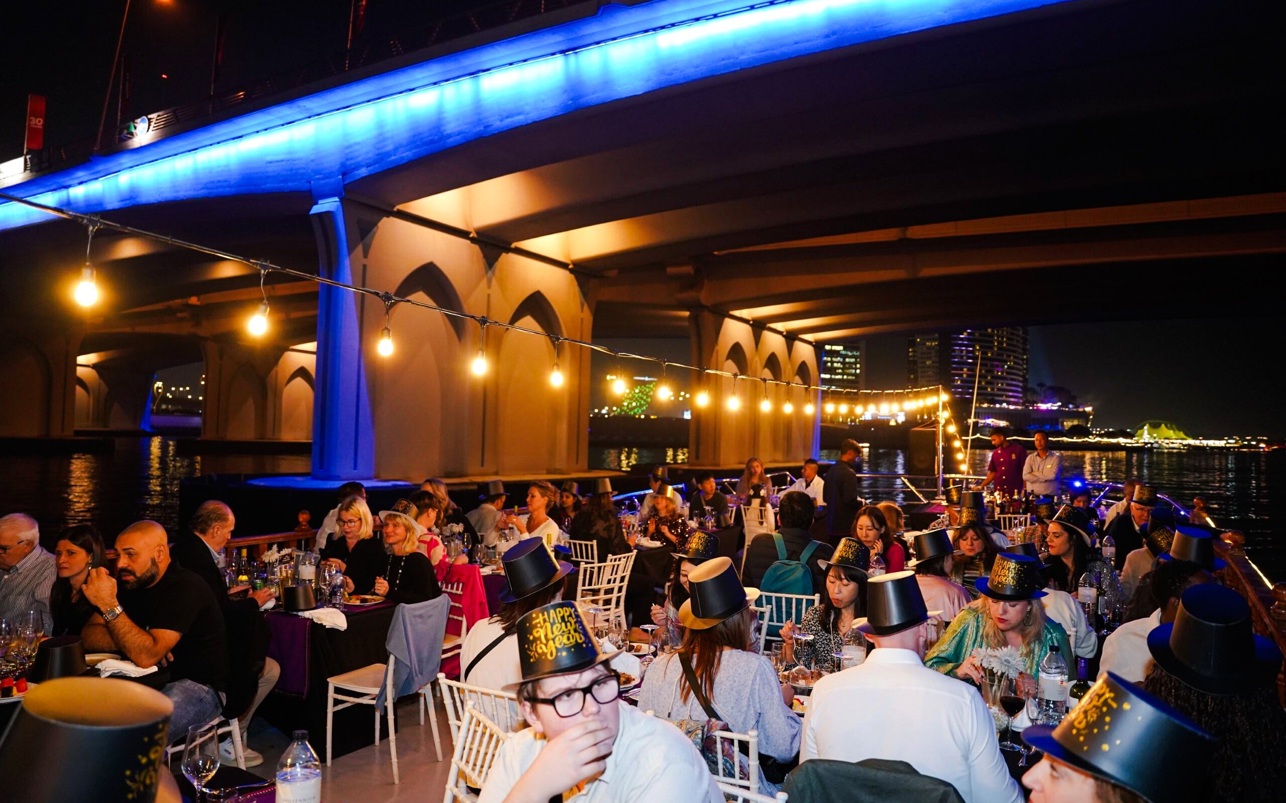 Guests dining on a New Year's Eve Creek Dhow Dinner Cruise under a lit bridge in Dubai.