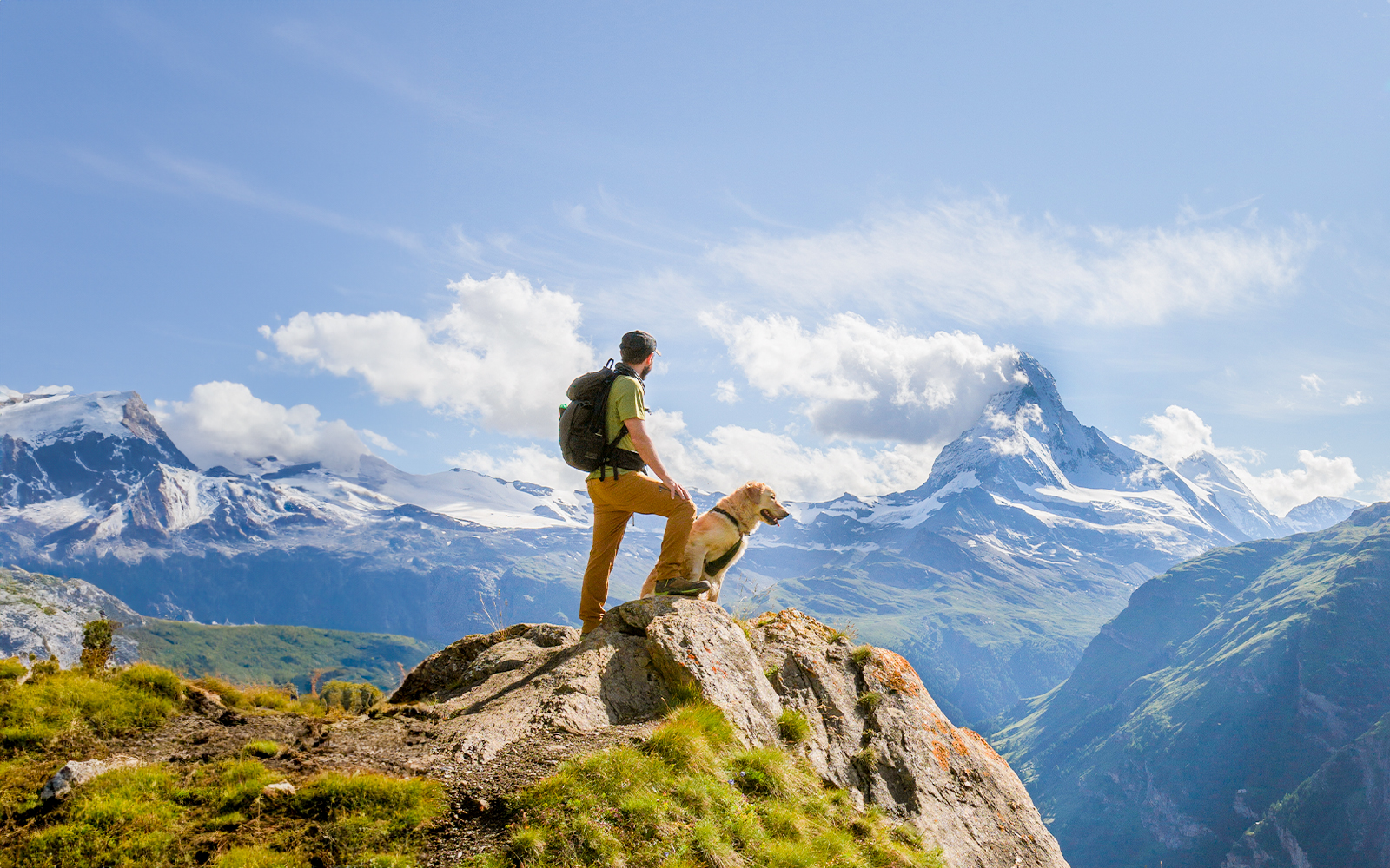 Man hiking with dog on mountain trail, overlooking snowy peaks in Switzerland.