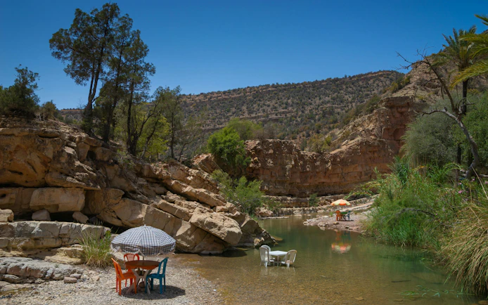 Chairs and tables by a river in Paradise Valley, surrounded by rocky cliffs and trees.