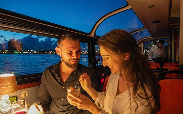 Couple enjoying dessert on an Amsterdam dinner cruise with city skyline in the background.