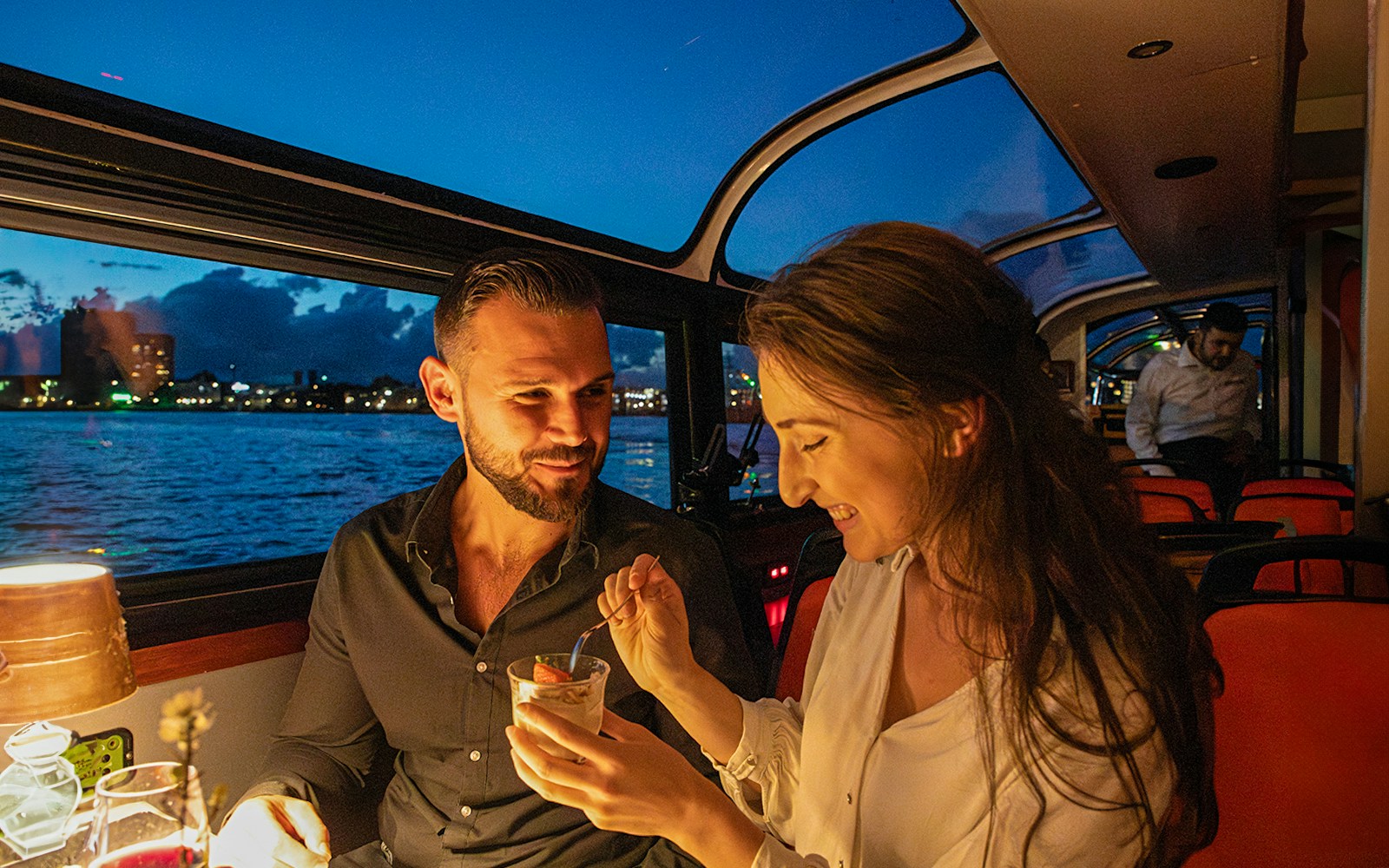 Couple enjoying dessert on an Amsterdam dinner cruise with city skyline in the background.
