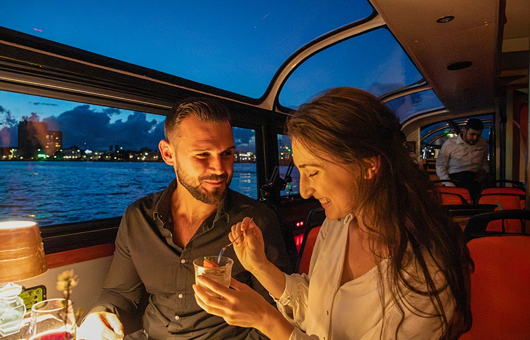 Couple enjoying dessert on an Amsterdam dinner cruise with city skyline in the background.