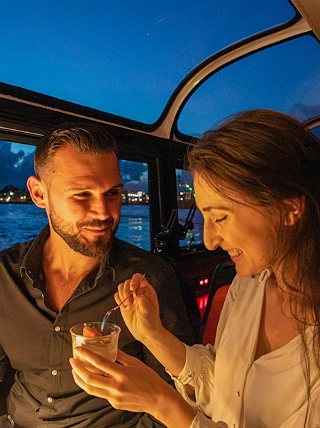Couple enjoying dessert on an Amsterdam dinner cruise with city skyline in the background.