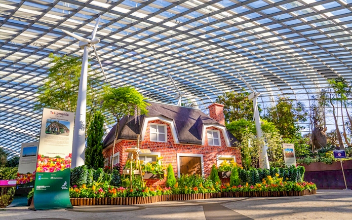 Brick house surrounded by tulips and greenery inside a glass conservatory.