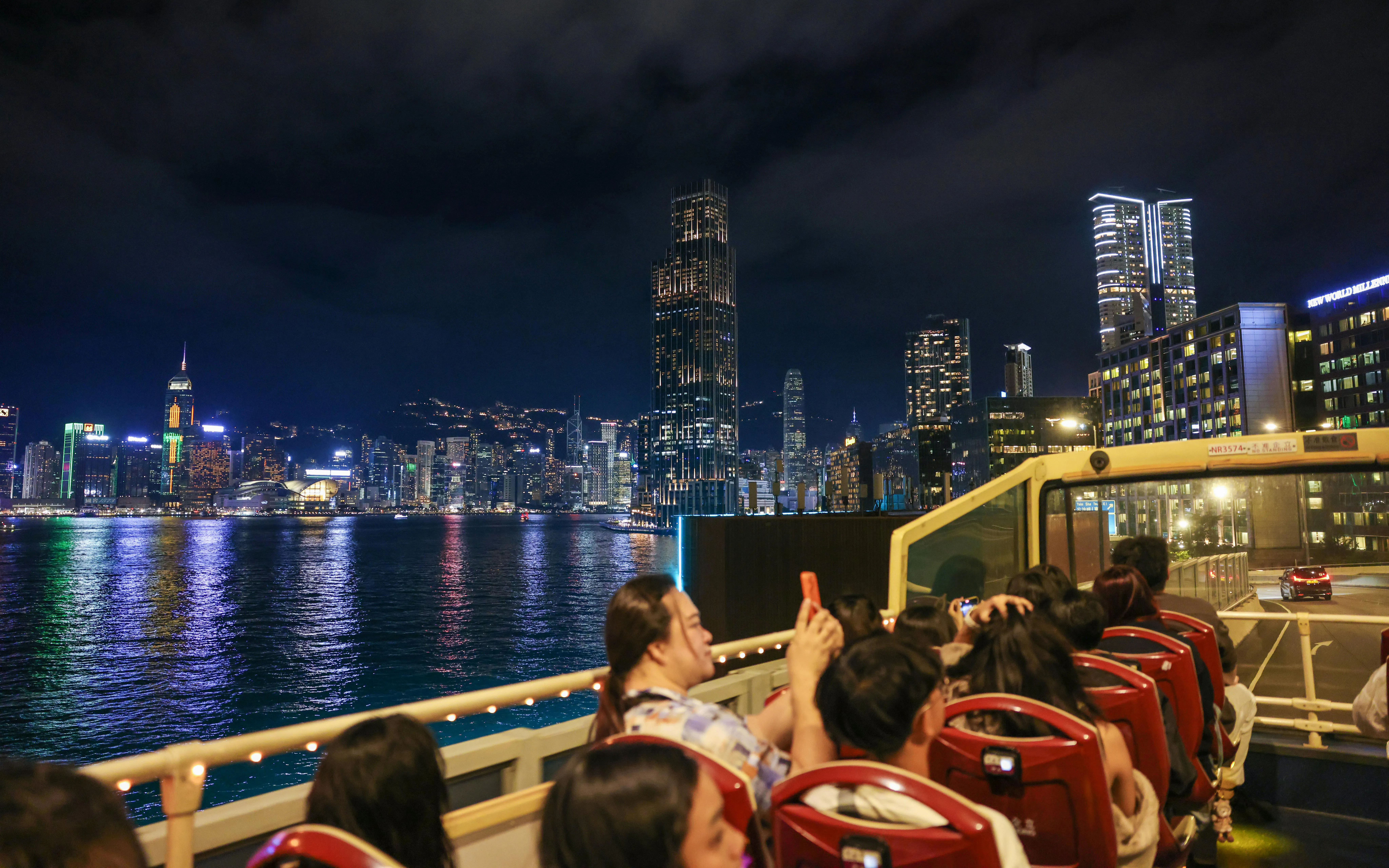 Open-top bus tour with passengers viewing Hong Kong skyline at night.