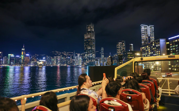 Open-top bus tour with passengers viewing Hong Kong skyline at night.