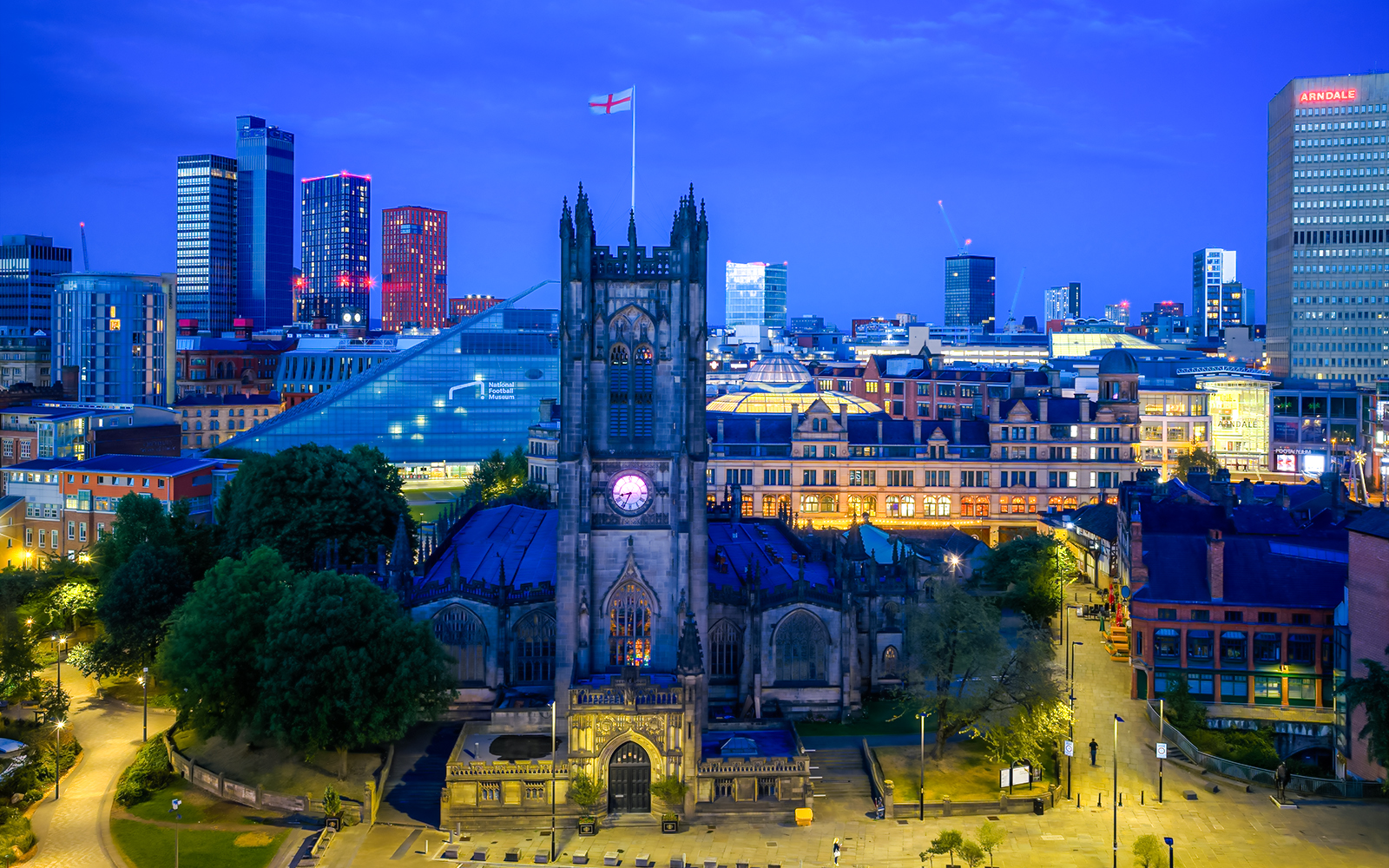 Manchester Cathedral exterior at dusk with city skyline in the background.