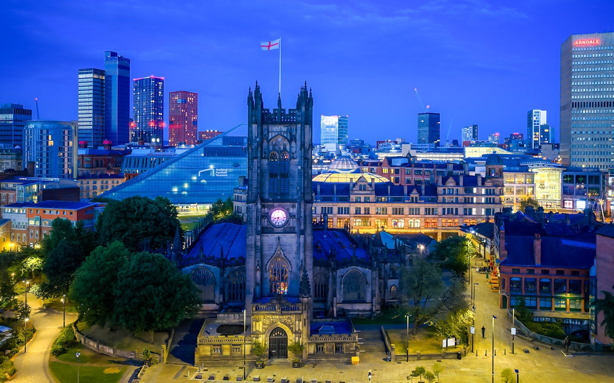 Manchester Cathedral exterior at dusk with city skyline in the background.