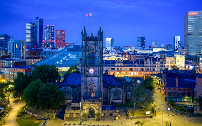 Manchester Cathedral exterior at dusk with city skyline in the background.