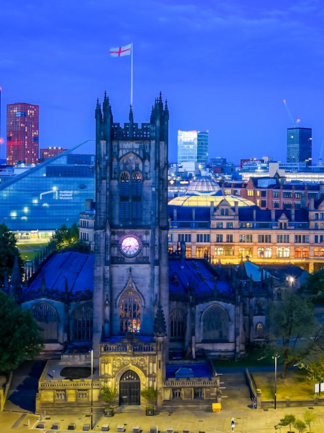 Manchester Cathedral exterior at dusk with city skyline in the background.