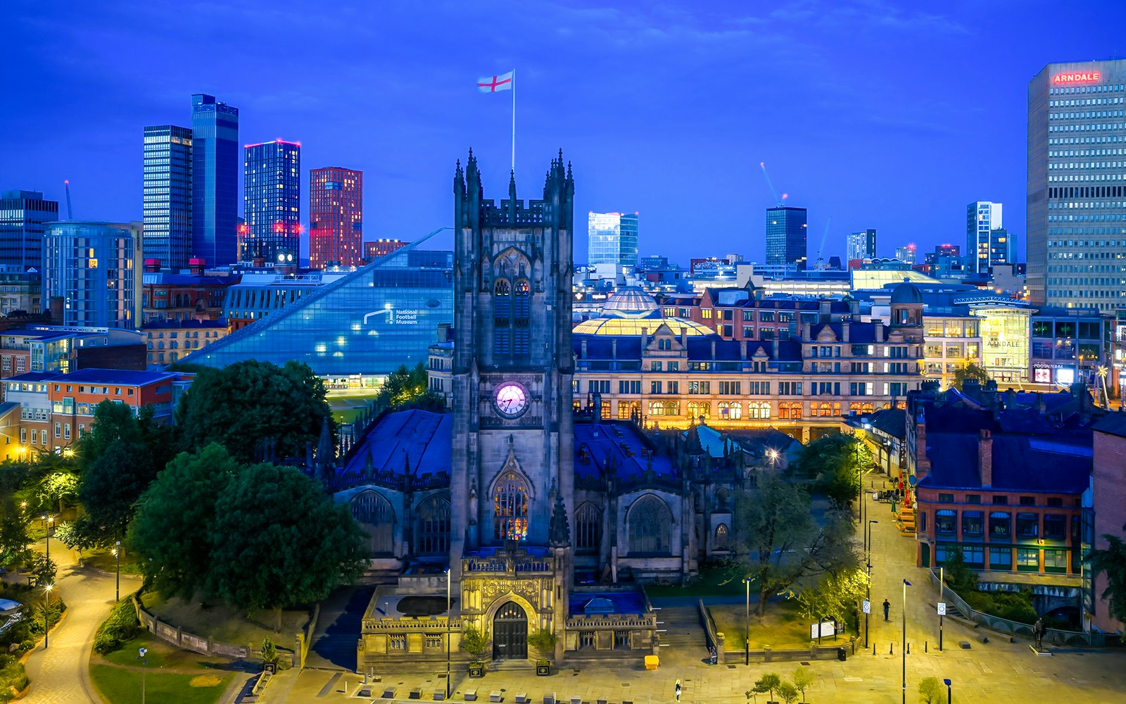 Manchester Cathedral exterior at dusk with city skyline in the background.