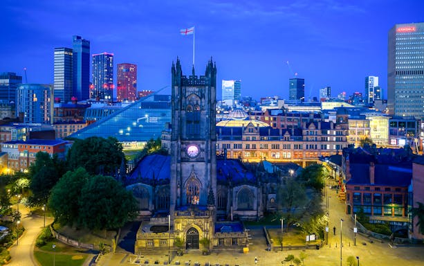 Manchester Cathedral exterior at dusk with city skyline in the background.