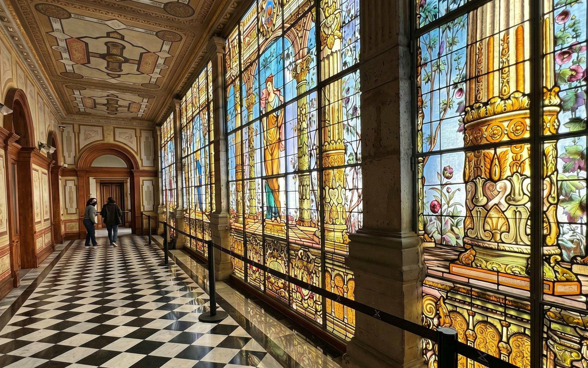 Stained glass corridor inside Chapultepec Castle, Mexico City, with intricate designs and visitors walking.