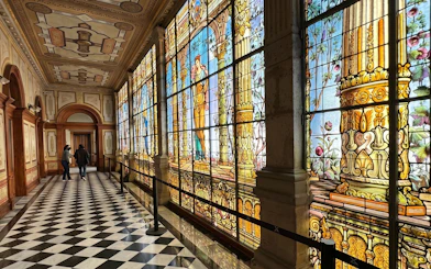 Stained glass corridor inside Chapultepec Castle, Mexico City, with intricate designs and visitors walking.