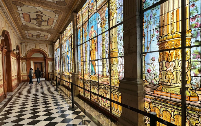 Stained glass corridor inside Chapultepec Castle, Mexico City, with intricate designs and visitors walking.