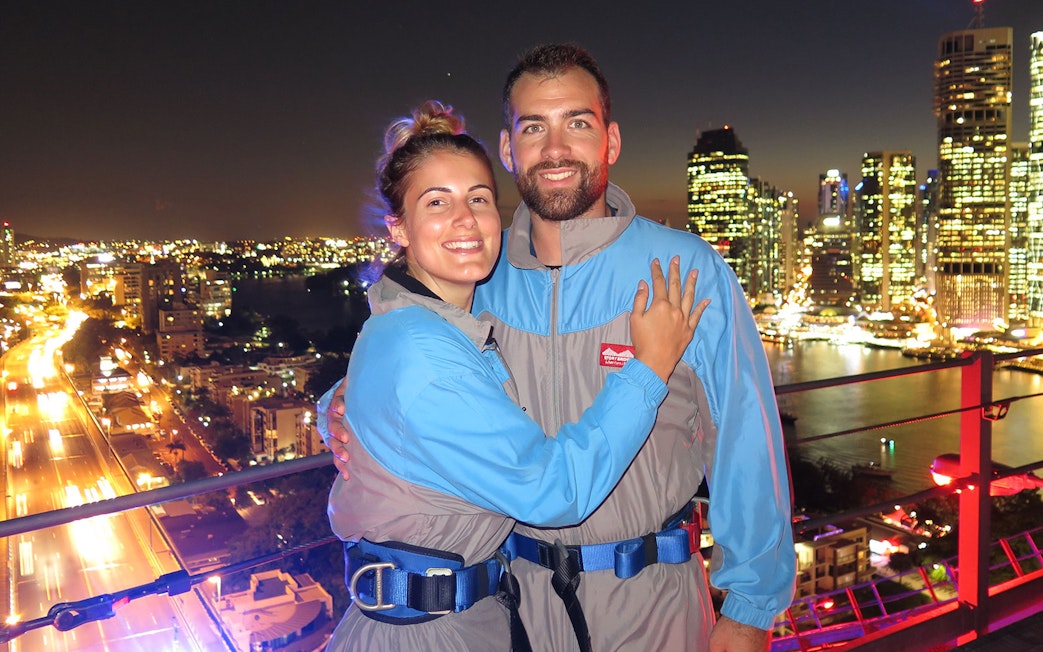 Couple on Brisbane Story Bridge during twilight climb with city lights in background.