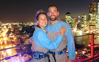 Couple on Brisbane Story Bridge during twilight climb with city lights in background.