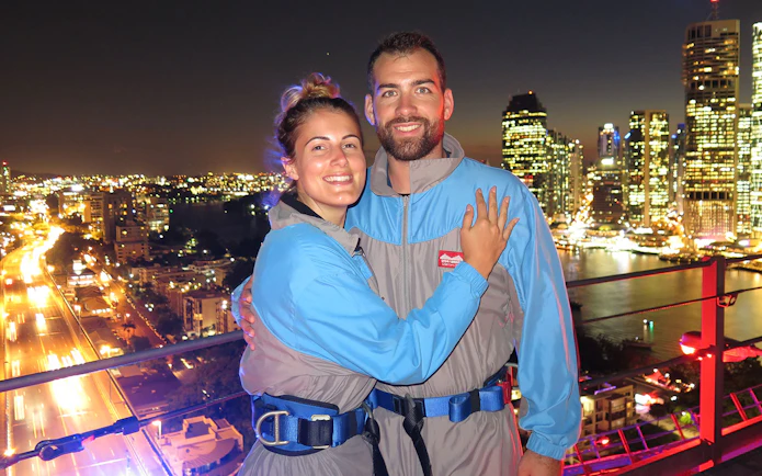 Couple on Brisbane Story Bridge during twilight climb with city lights in background.