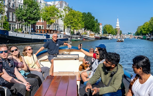 Small open boat with tourists cruising Amsterdam canal, view of Montelbaanstoren tower.
