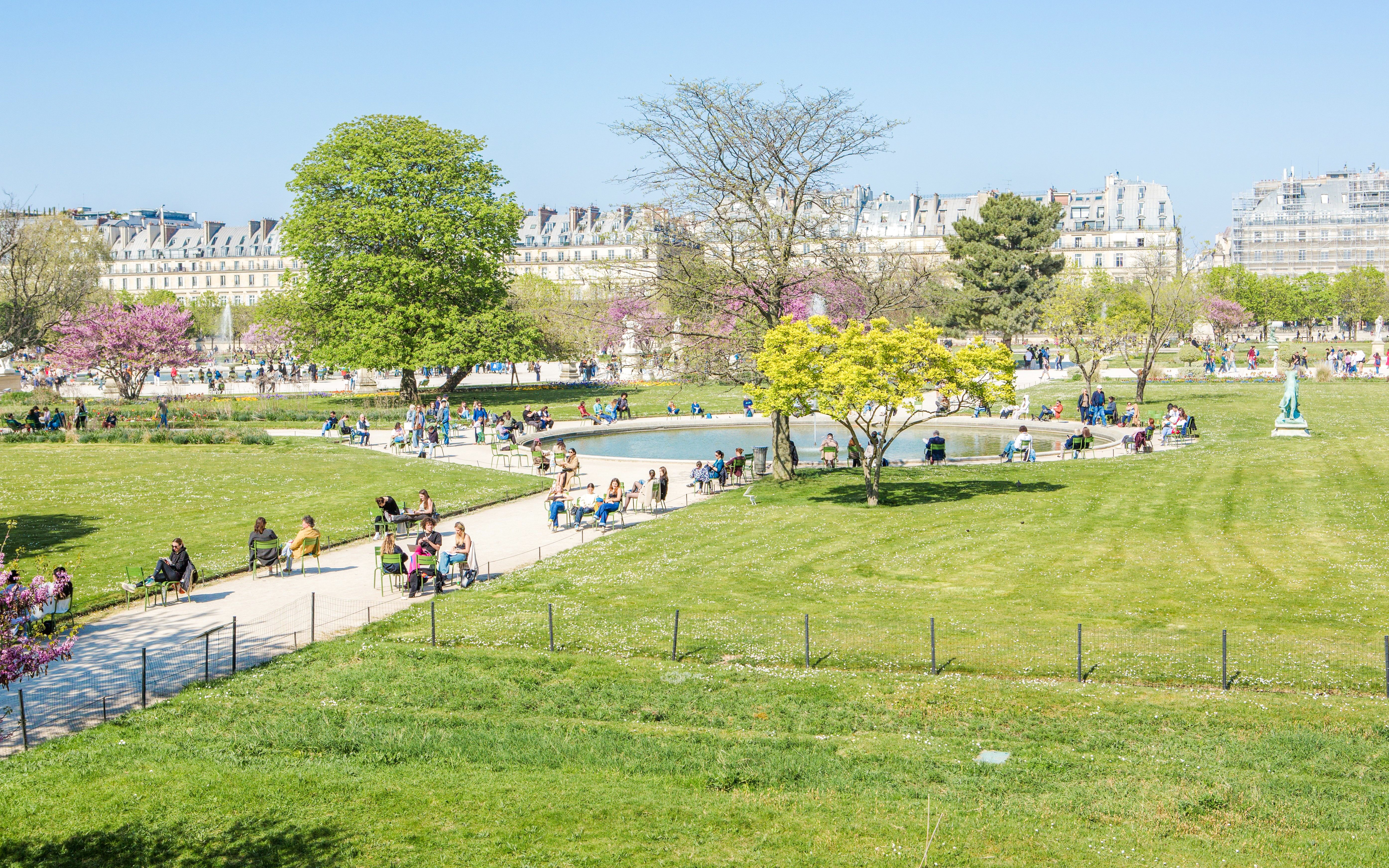 Visitors relaxing by the pond in Tuileries Garden, Paris, with trees and historic buildings in the background.