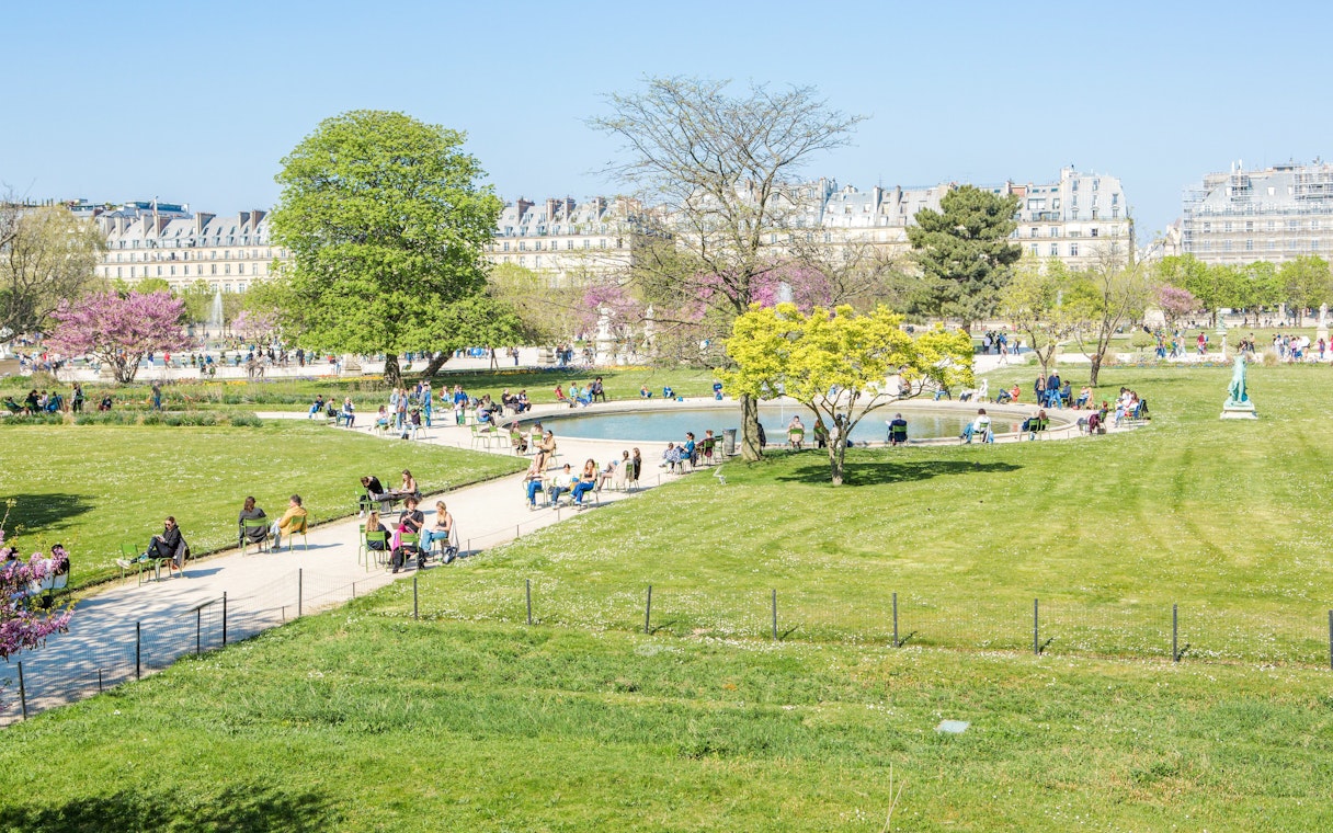 Visitors relaxing by the pond in Tuileries Garden, Paris, with trees and historic buildings in the background.