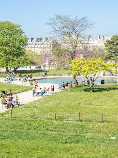 Visitors relaxing by the pond in Tuileries Garden, Paris, with trees and historic buildings in the background.