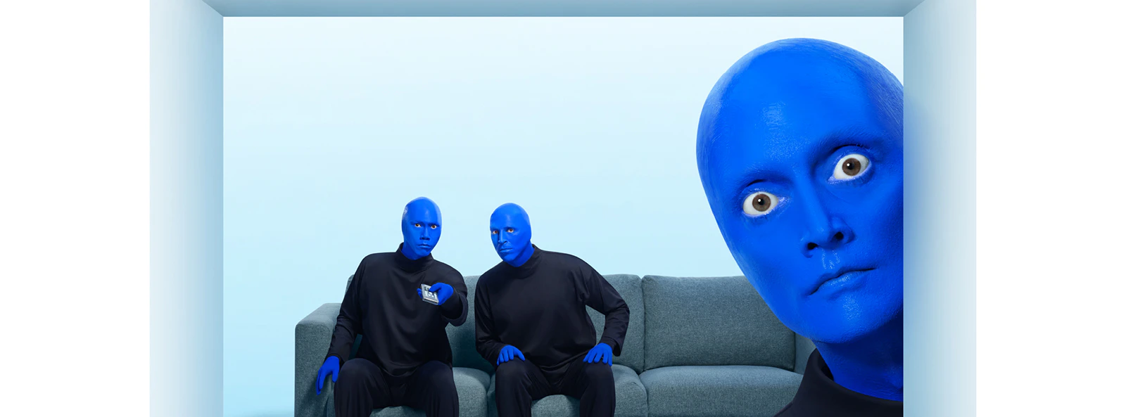 Blue Man Group members in blue makeup sitting on a couch with one peeking from the side.