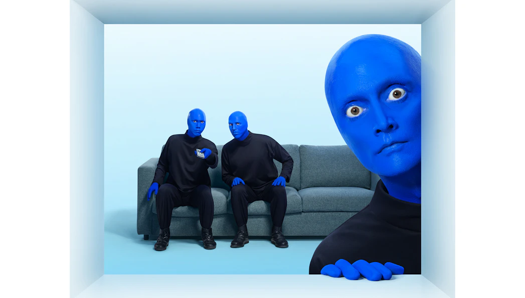 Blue Man Group members in blue makeup sitting on a couch with one peeking from the side.