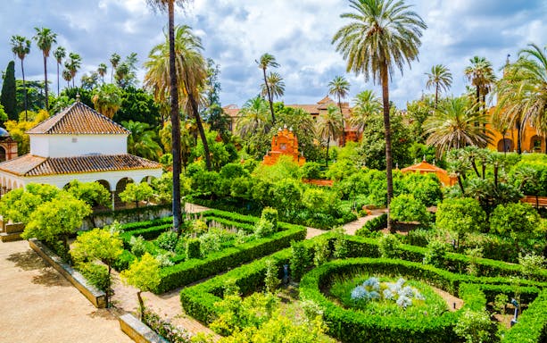 Alcazar Seville gardens with lush greenery, palm trees, and historic architecture.