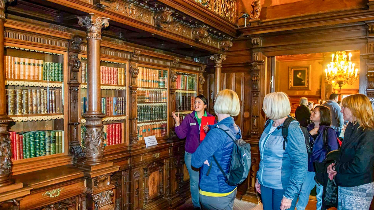 Guided tour group in Peles Castle library, Romania, viewing ornate wooden bookshelves.