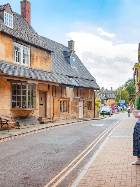 Cotswolds street scene with historic buildings and a person taking photos.
