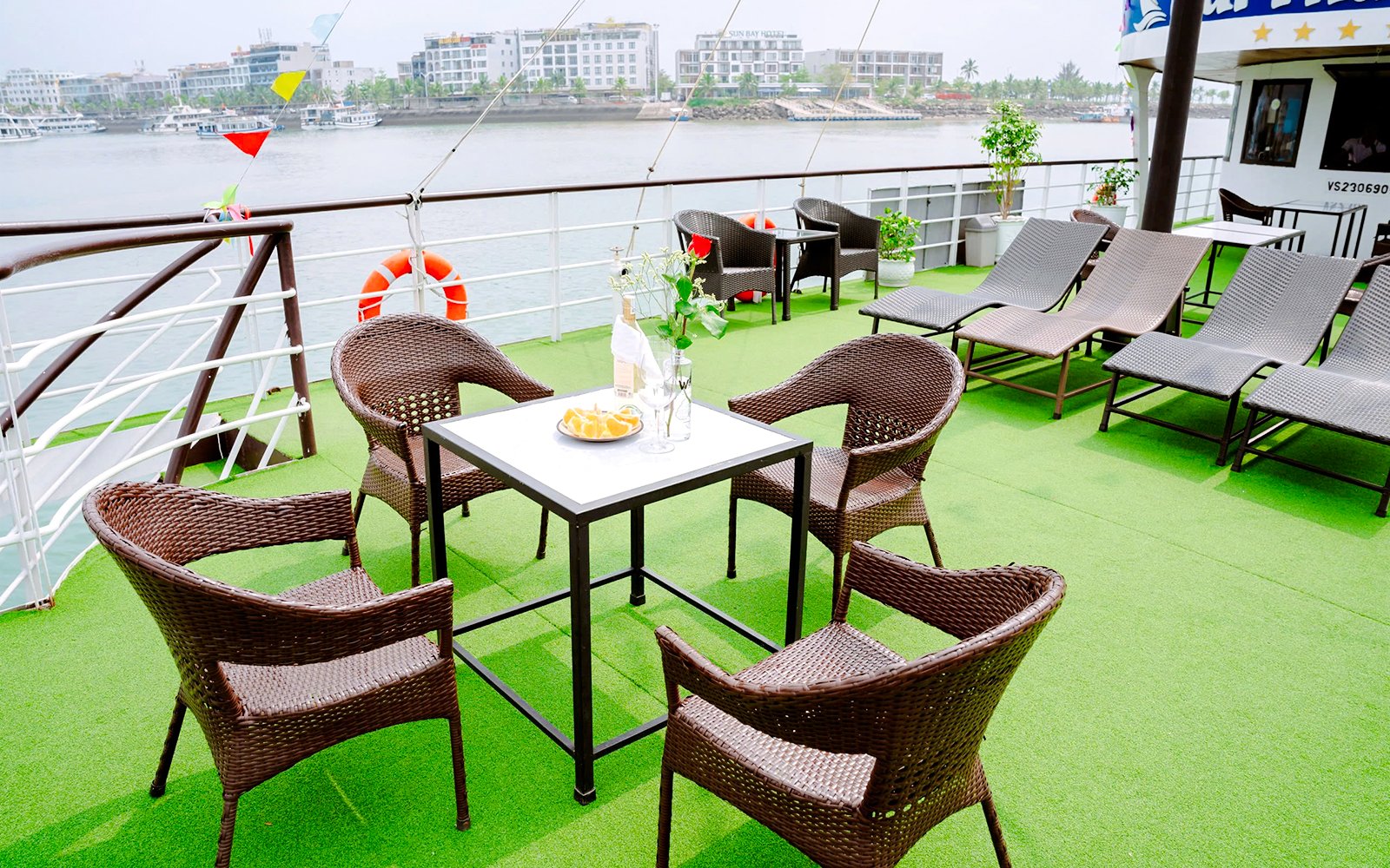 Outdoor seating area on cruise ship deck with wicker chairs and table, overlooking water.