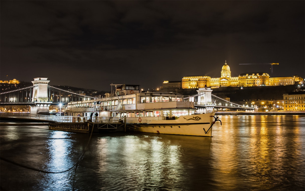 Sightseeing cruise on Danube River with Buda Castle and Chain Bridge illuminated at night.