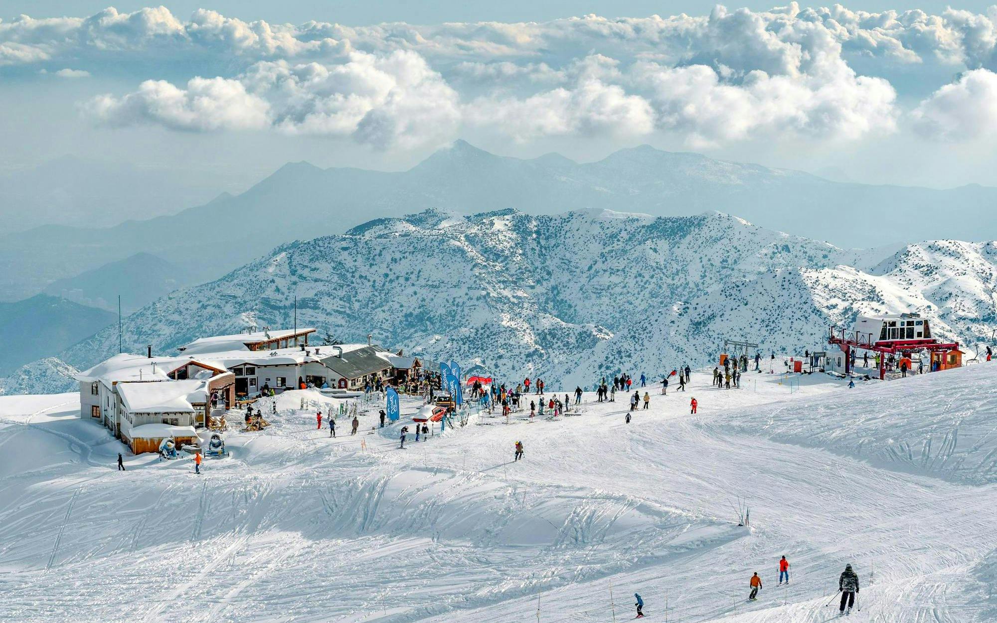 Tourists skiing and snowboarding at Farellones Park with snowy mountains in the background.