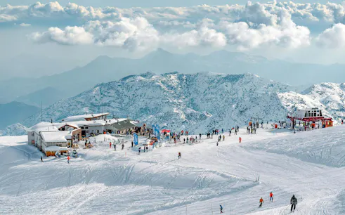 Tourists skiing and snowboarding at Farellones Park with snowy mountains in the background.
