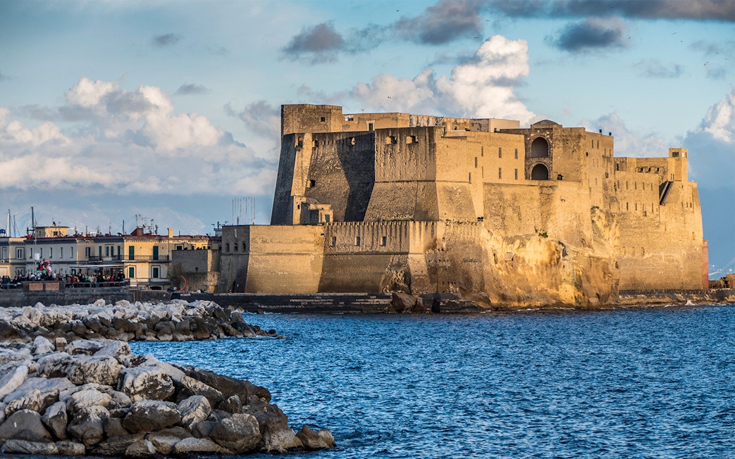 Castel dell'Ovo on the Naples waterfront with rocky shoreline and blue sky.