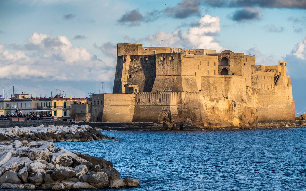 Castel dell'Ovo on the Naples waterfront with rocky shoreline and blue sky.