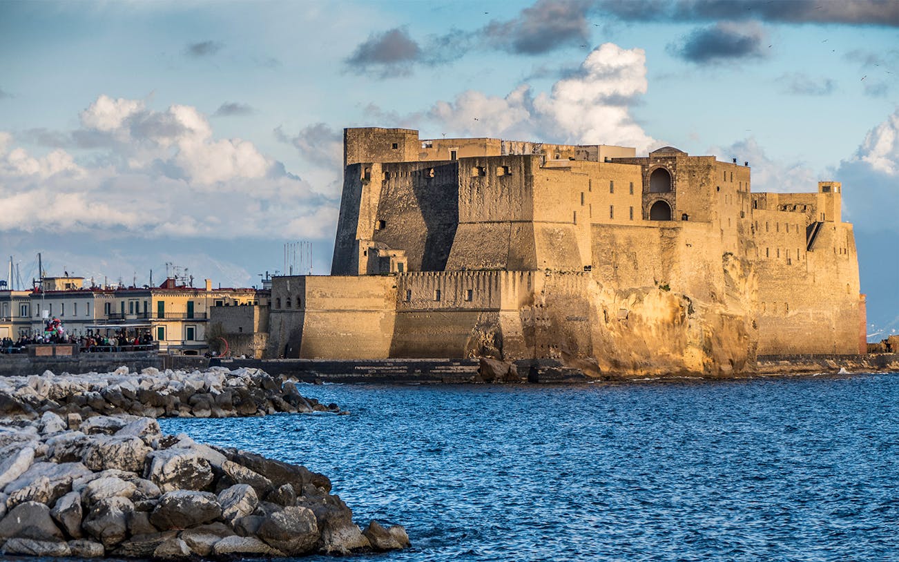 Castel dell'Ovo on the Naples waterfront with rocky shoreline and blue sky.