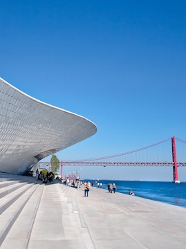 MAAT museum exterior with visitors and Lisbon's 25 de Abril Bridge in the background.
