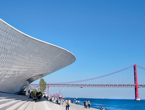 MAAT museum exterior with visitors and Lisbon's 25 de Abril Bridge in the background.