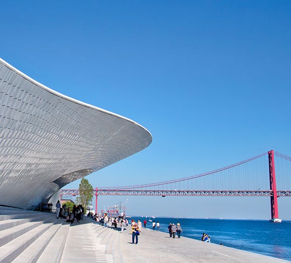 MAAT museum exterior with visitors and Lisbon's 25 de Abril Bridge in the background.