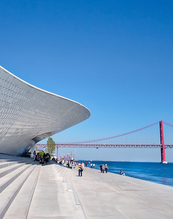 MAAT museum exterior with visitors and Lisbon's 25 de Abril Bridge in the background.
