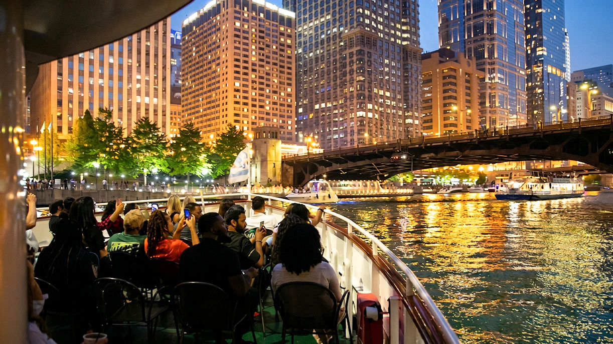 Cruise passengers on Chicago River at night with city skyline and bridge in view.